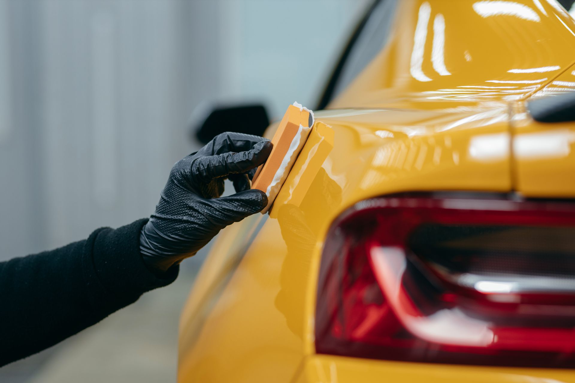 Gloved hand applying ceramic coating to a yellow car's rear quarter panel with a sponge-like applicator.