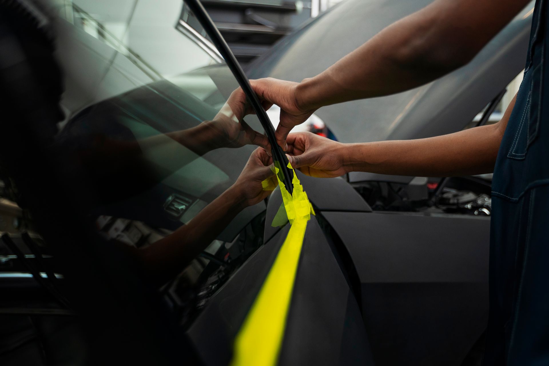 Hands applying yellow tape to a car's black body, near the windshield.