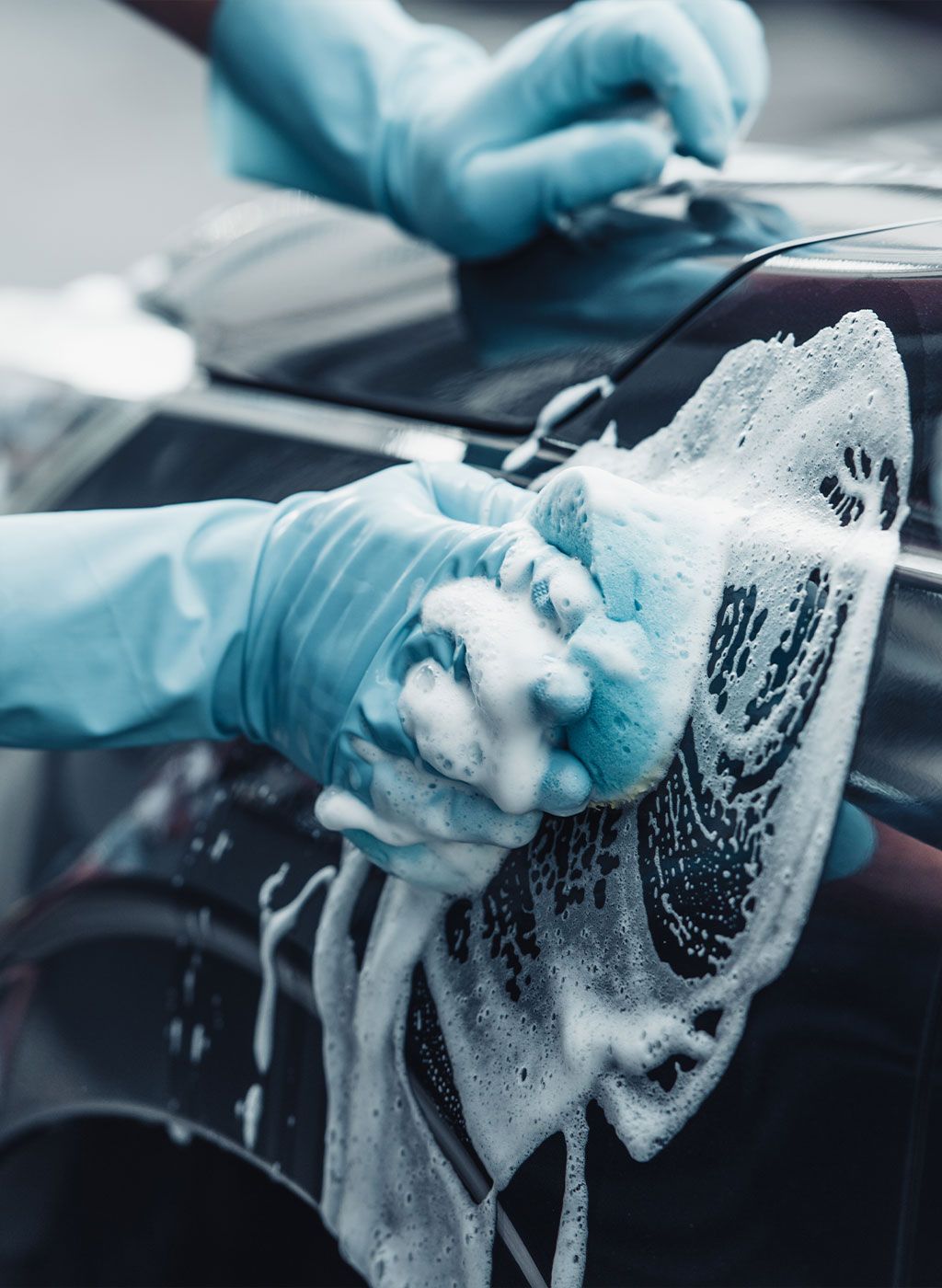 Hands in blue gloves washing a dark car with a soapy sponge.