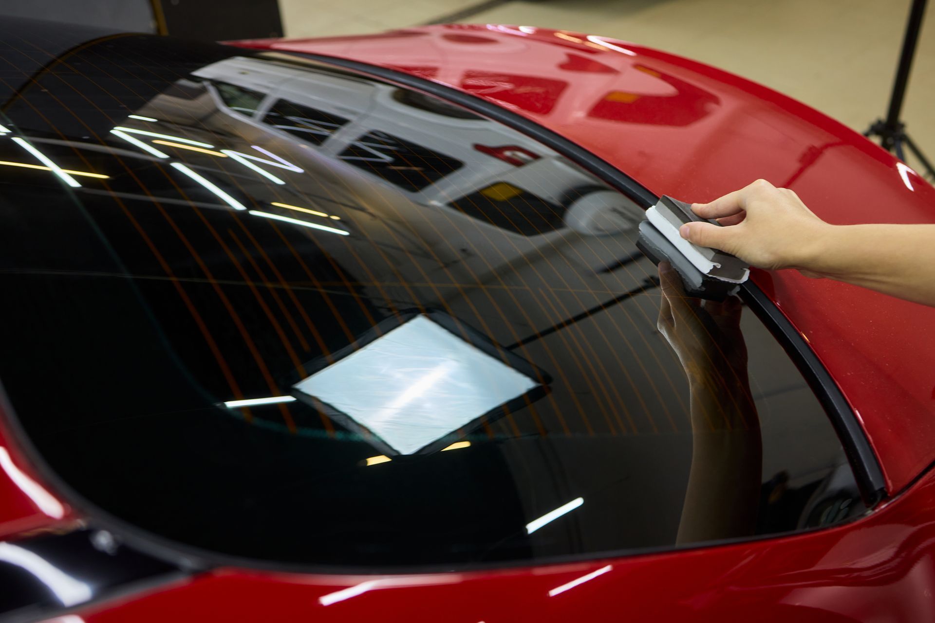 Person applying tint film to a red car's rear window with a tool.