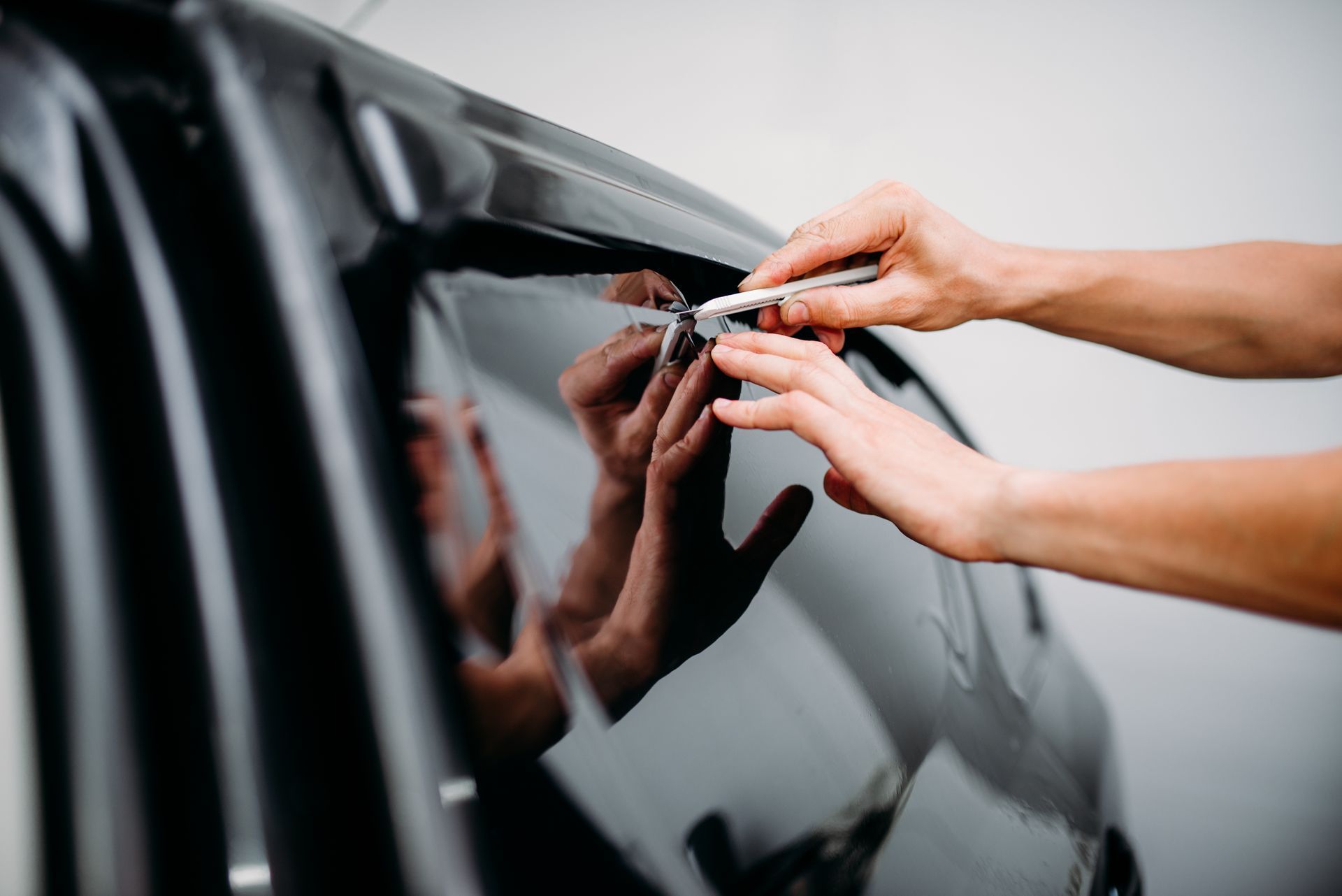 Hands using a utility knife to trim car window tinting film on a vehicle.
