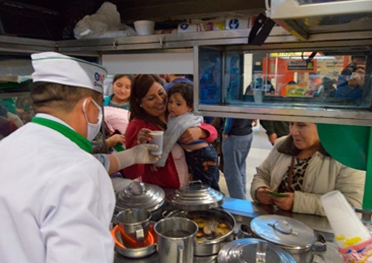 Un hombre con una máscara le sirve una taza de café a una mujer que sostiene a un bebé.