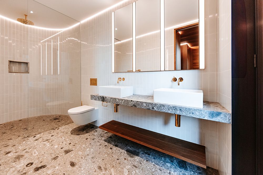 Modern bathroom with white sinks, terrazzo floor, and wall-mounted toilet. Gold fixtures and back-lit mirrors — BSpoke Cabinet Making & Joinery in Shellharbour, NSW