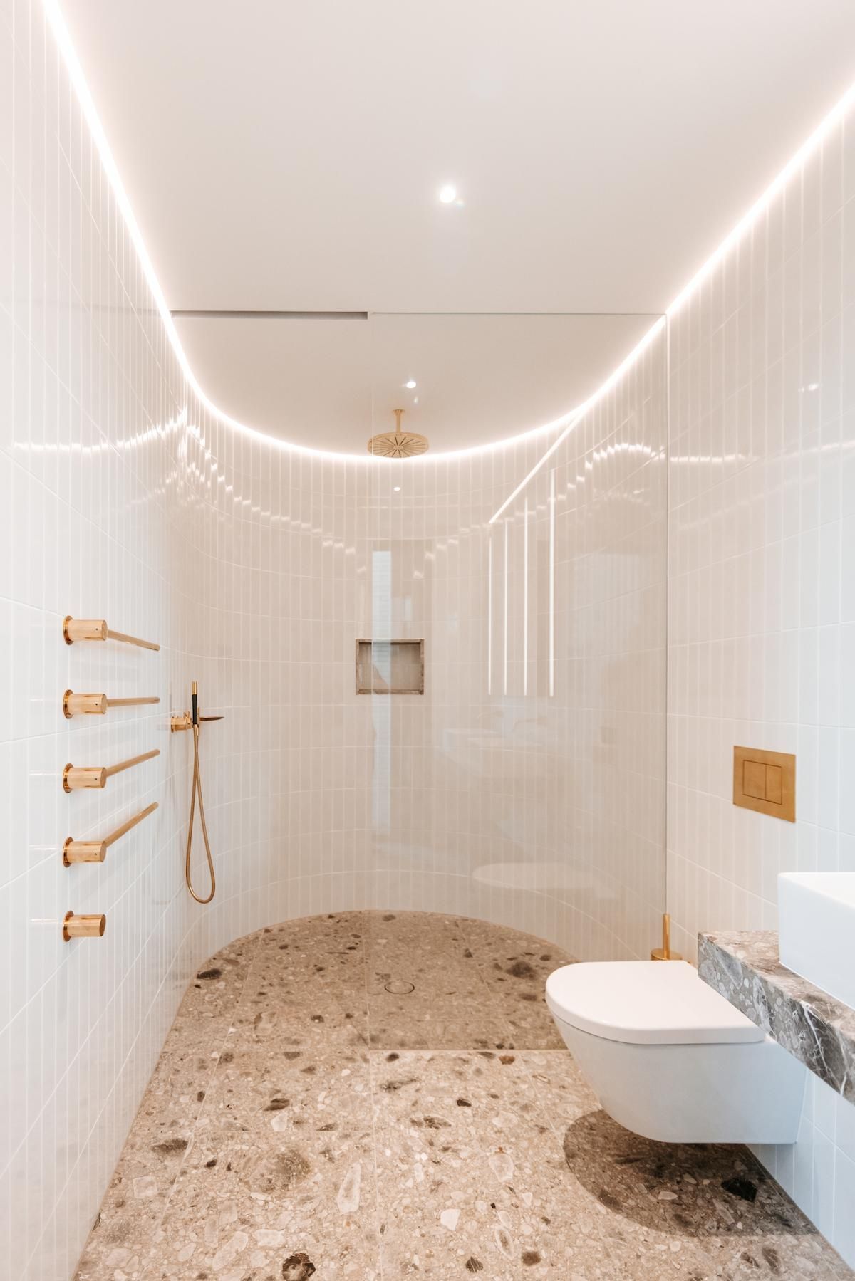 Modern white bathroom with gold fixtures, terrazzo floor, and curved ceiling lighting — BSpoke Cabinet Making & Joinery in Shellharbour, NSW