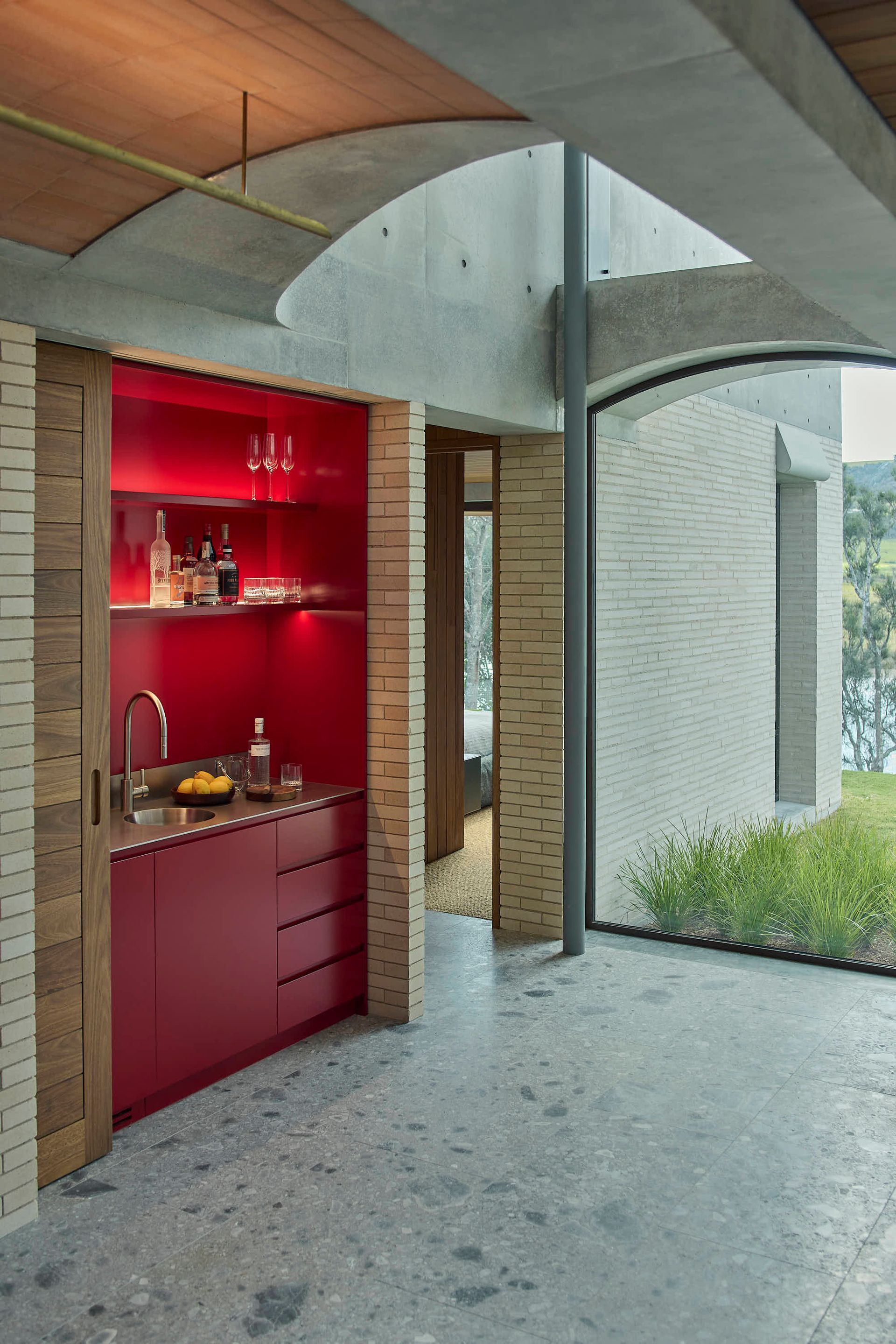 Red bar area with sink, shelves, and drinks, set against brick and curved glass — BSpoke Cabinet Making & Joinery in Unanderra, NSW