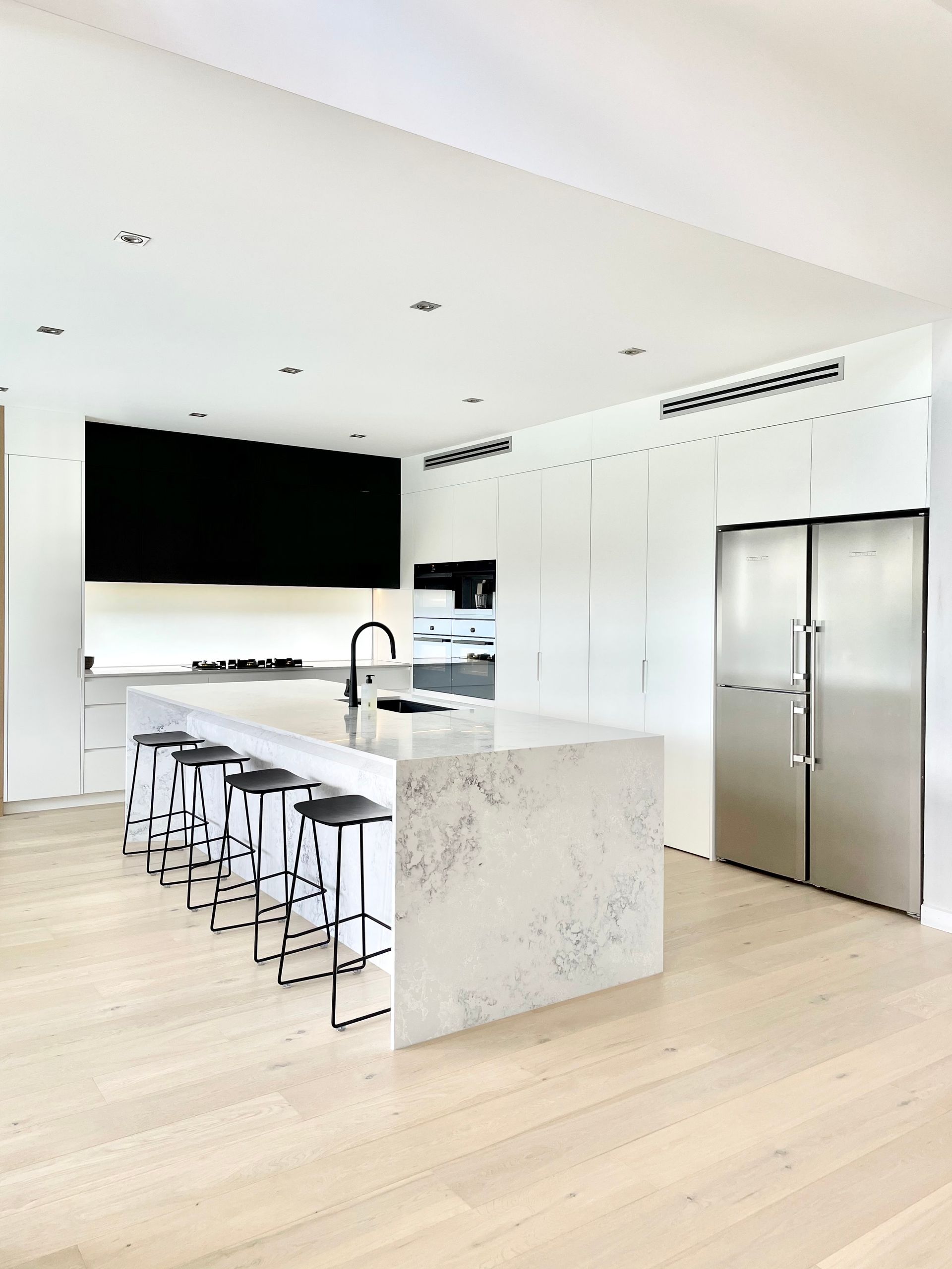 Modern kitchen with white cabinets, marble island with black stools, stainless steel refrigerator, and light wood floor — BSpoke Cabinet Making & Joinery in Unanderra, NSW