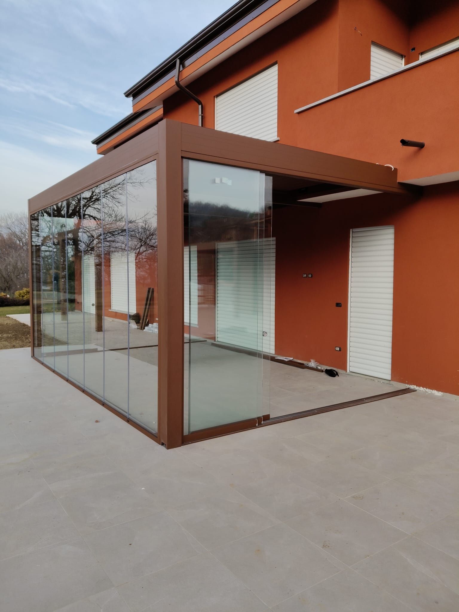 Veranda con pareti in vetro annessa a una casa color ruggine, struttura marrone, patio in cemento e persiane bianche.