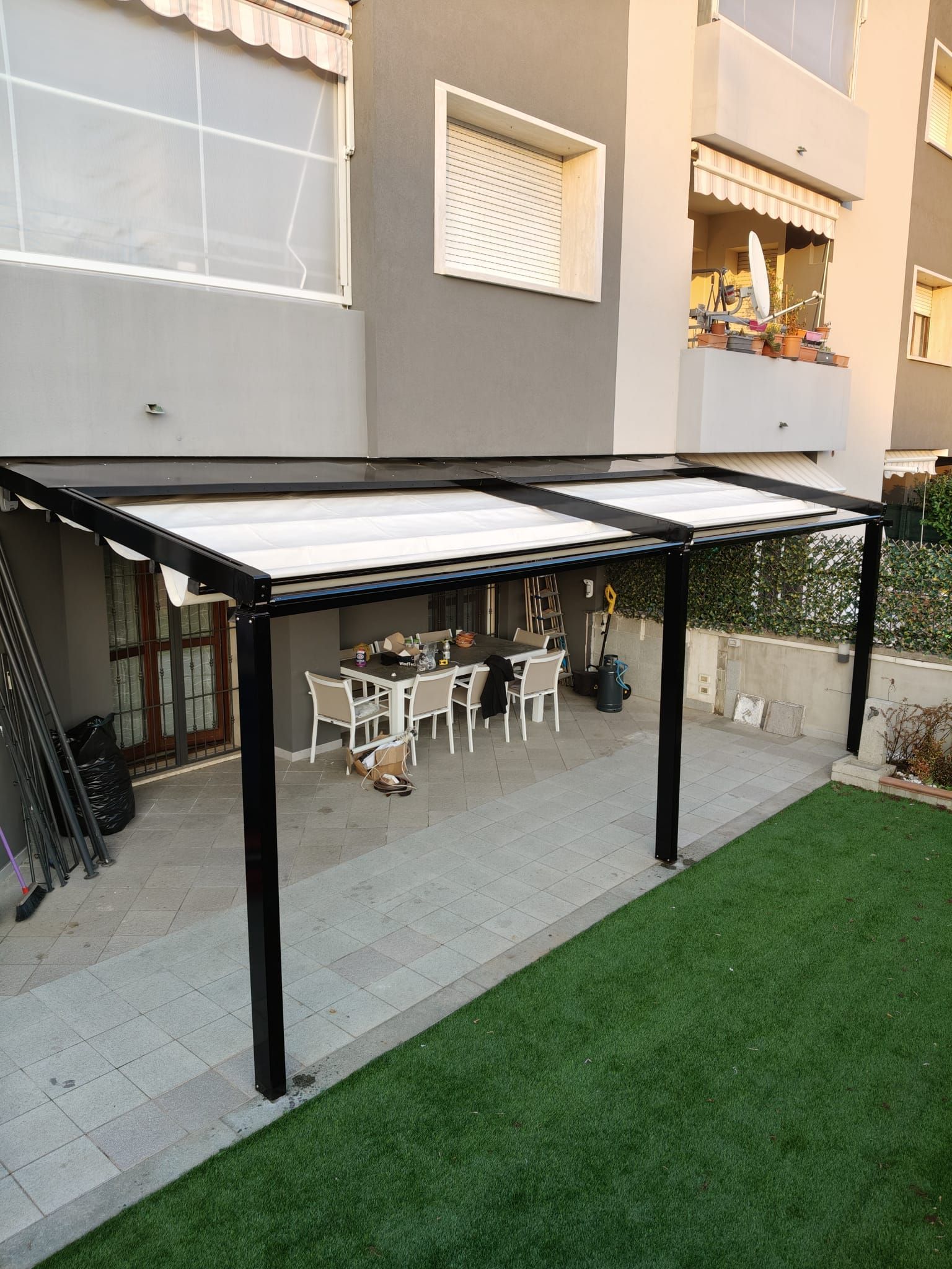 Pergola nera sopra il patio con tavolo da pranzo. Prato verde e un edificio grigio chiaro sullo sfondo.