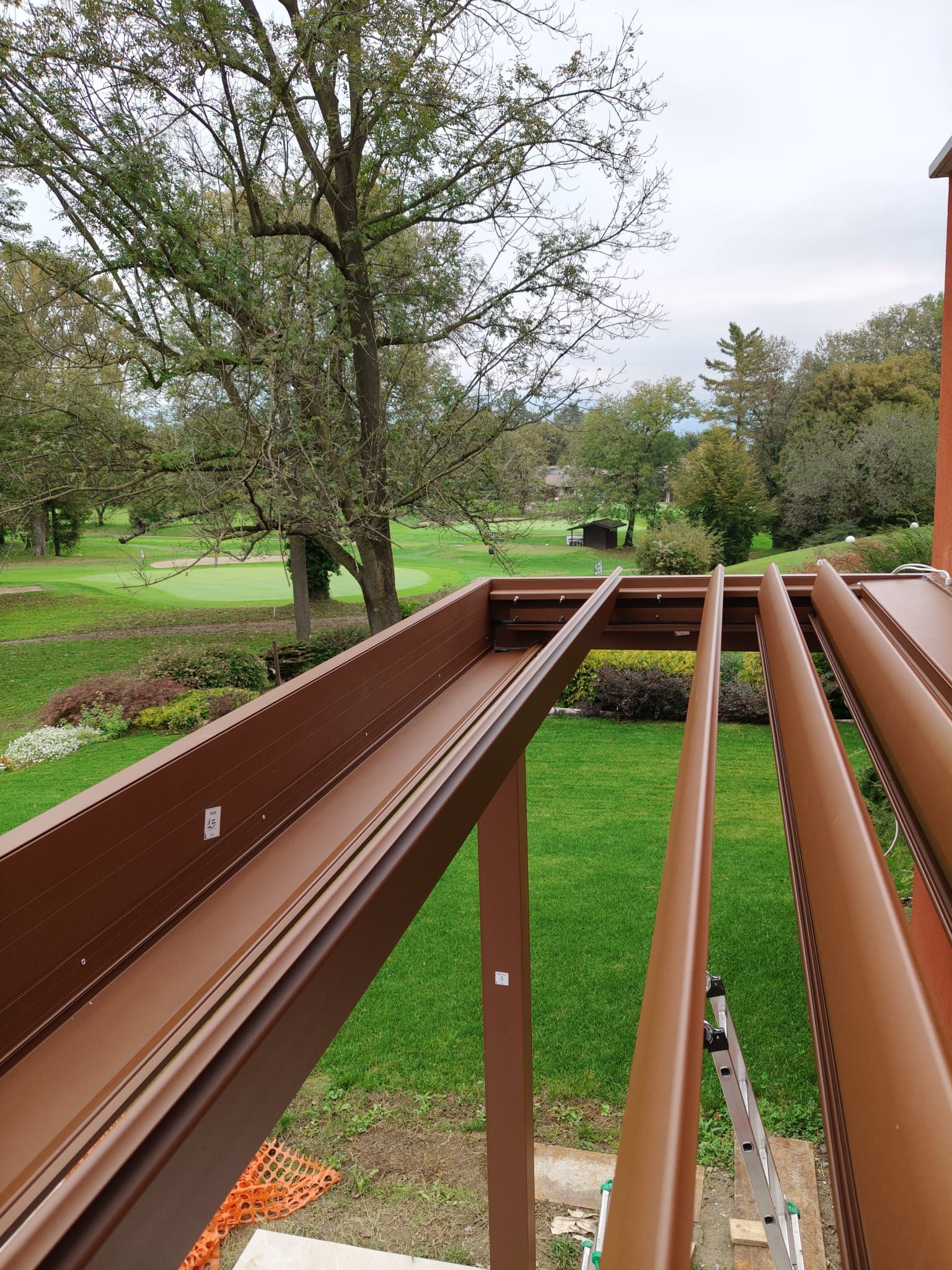 Pergola marrone con persiane aperte che si affaccia su un cortile verde e sugli alberi.