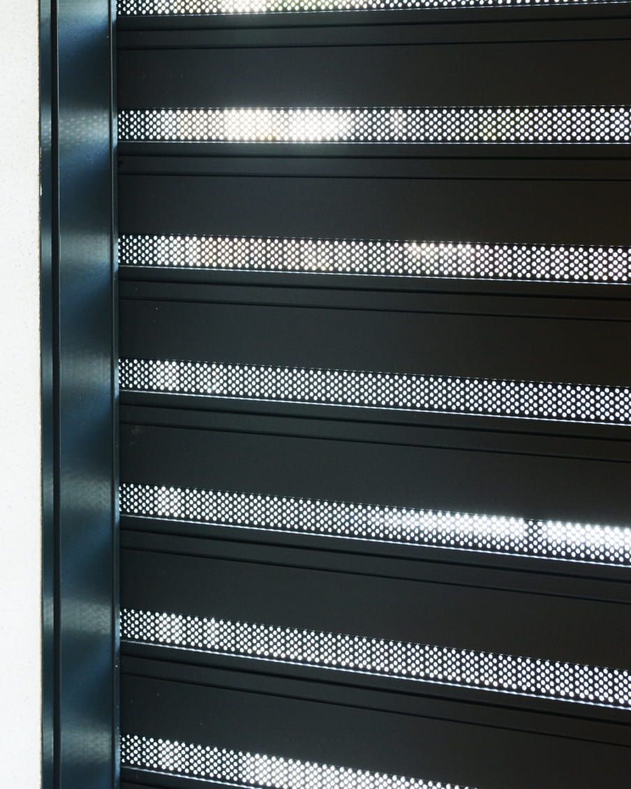 Primo piano di tende perforate grigio scuro che lasciano passare raggi di luce solare.