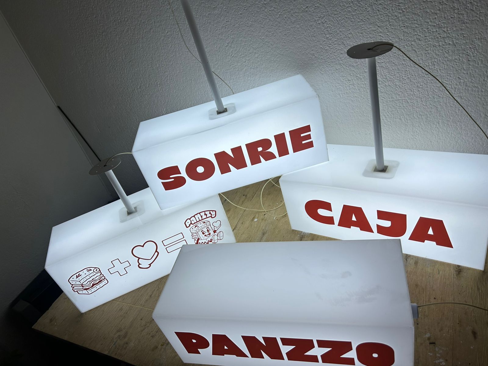 Cuatro letreros blancos iluminados colgados del techo, cada uno con texto rojo. Las palabras son 