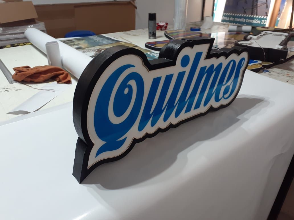 Letrero con el logo de cerveza Quilmes en color azul y blanco con borde negro sobre superficie blanca.