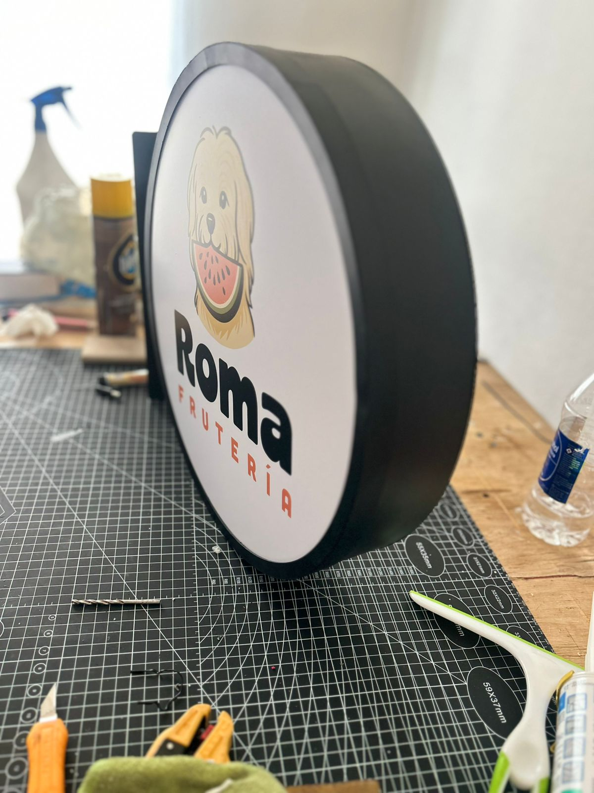 Letrero redondo de frutería Roma con un perro comiendo sandía, marco negro, sobre tapete de corte.