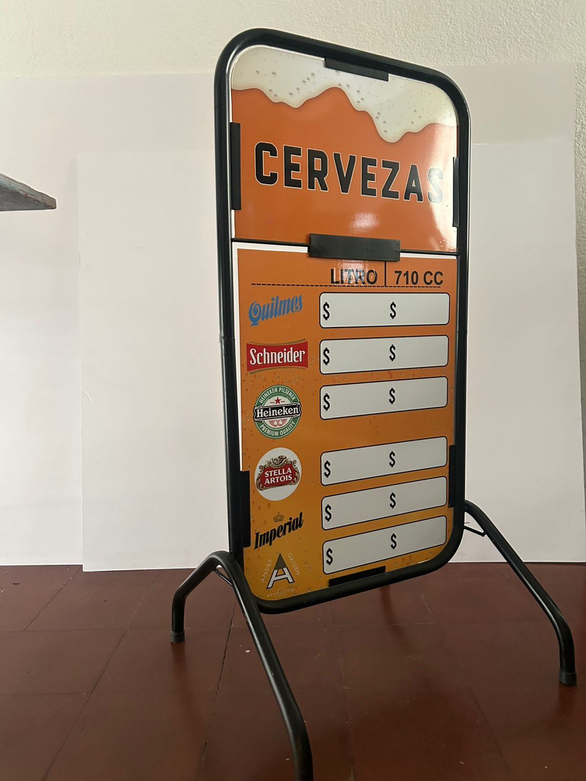 Letrero publicitario de cerveza con marco en forma de A; incluye precios y marcas. Fondo naranja y marco negro.