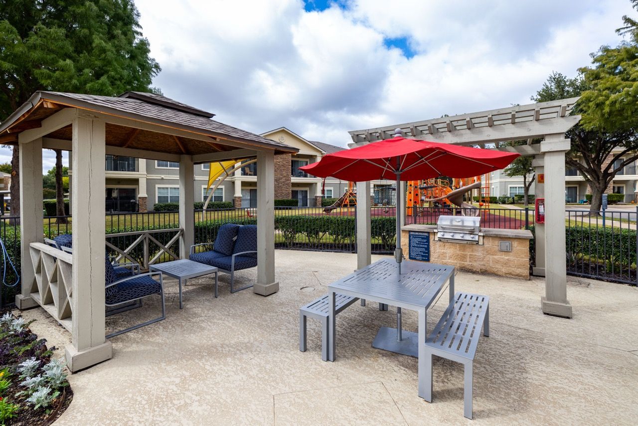 Outdoor community patio with seating under a pergola, a grill, and a red umbrella.