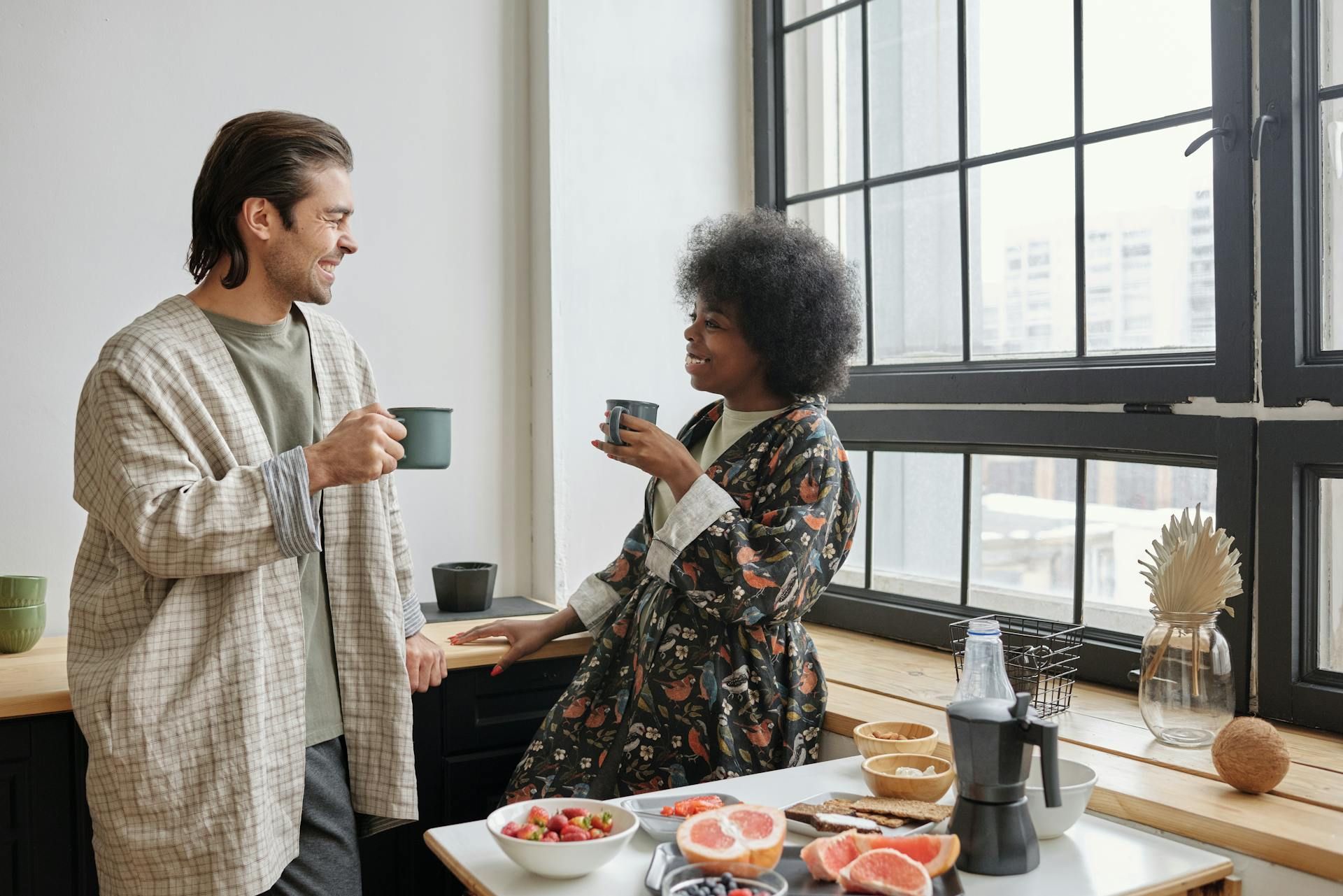 Two people in robes stand by a window in a bright kitchen, talking and holding mugs while fruit sits on the table.