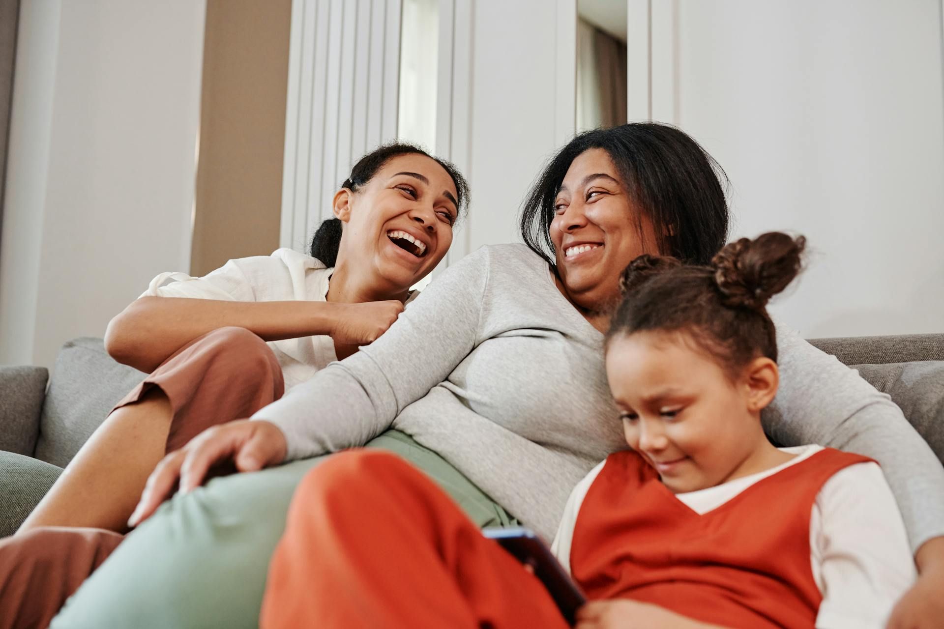 Three people sit together on a sofa, laughing and looking down at a phone held by the person in the foreground.