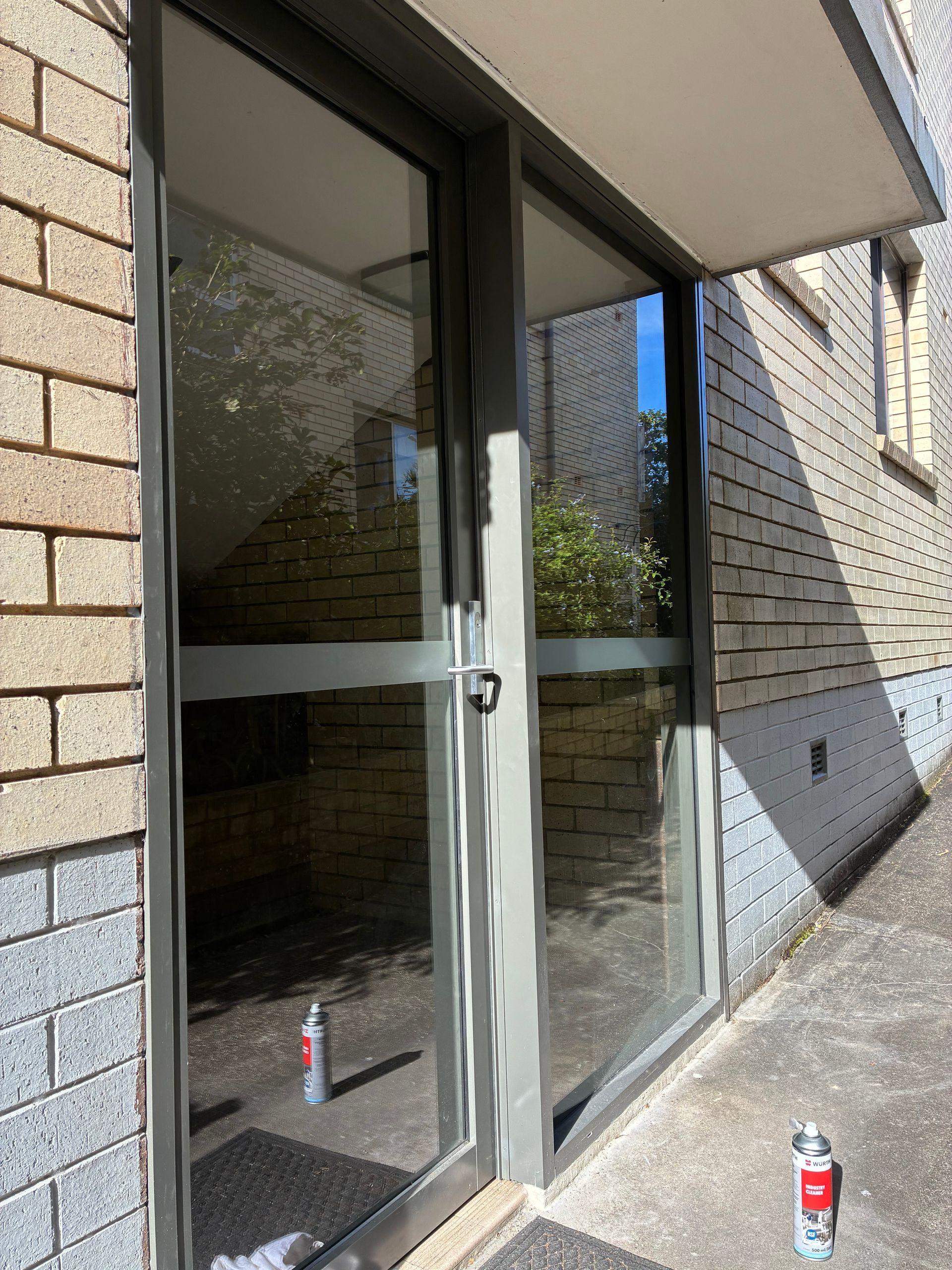 Glass entrance doors beside a brick building, with soda cans on the ground outside. — Kracken Windows and Doors In Terrey Hills, NSW