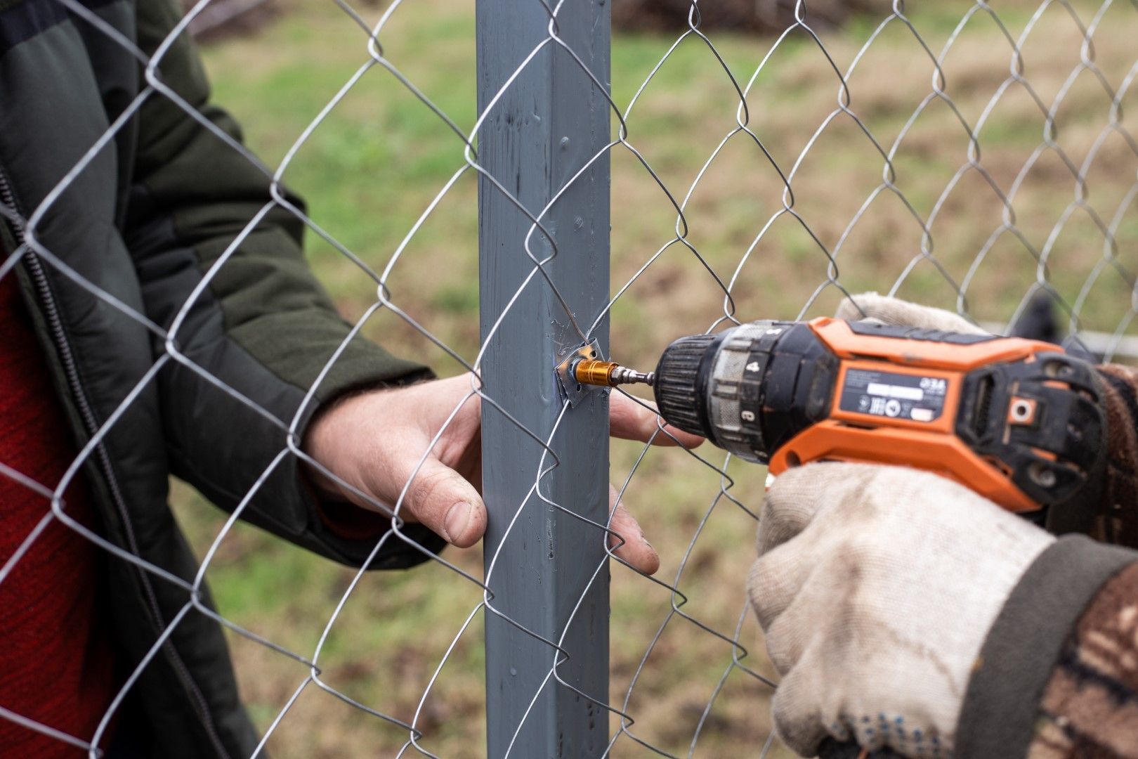 Person using a power drill to attach chain-link fence to a gray post outdoors.
