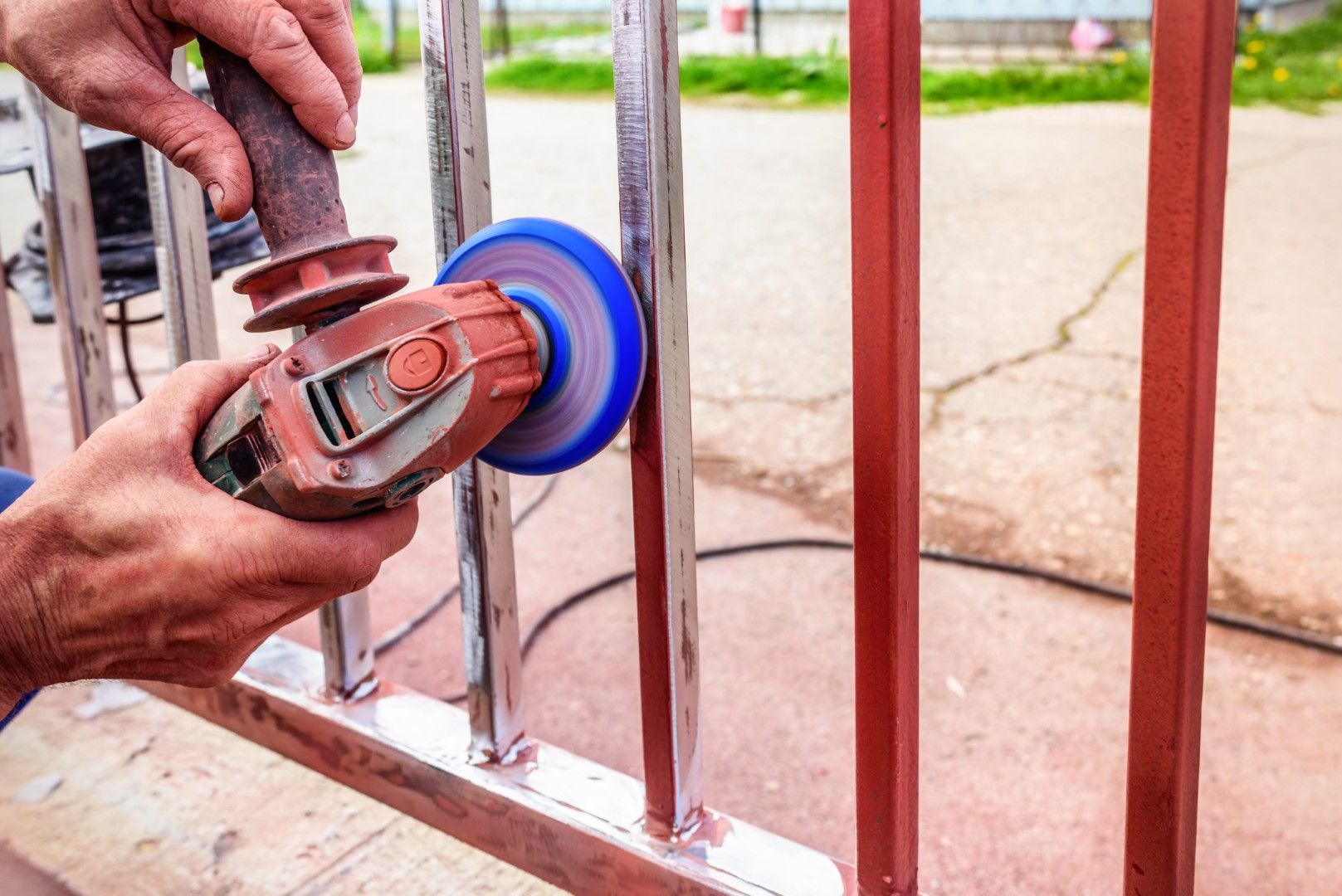 Person using an angle grinder to remove paint from a red metal fence outdoors.