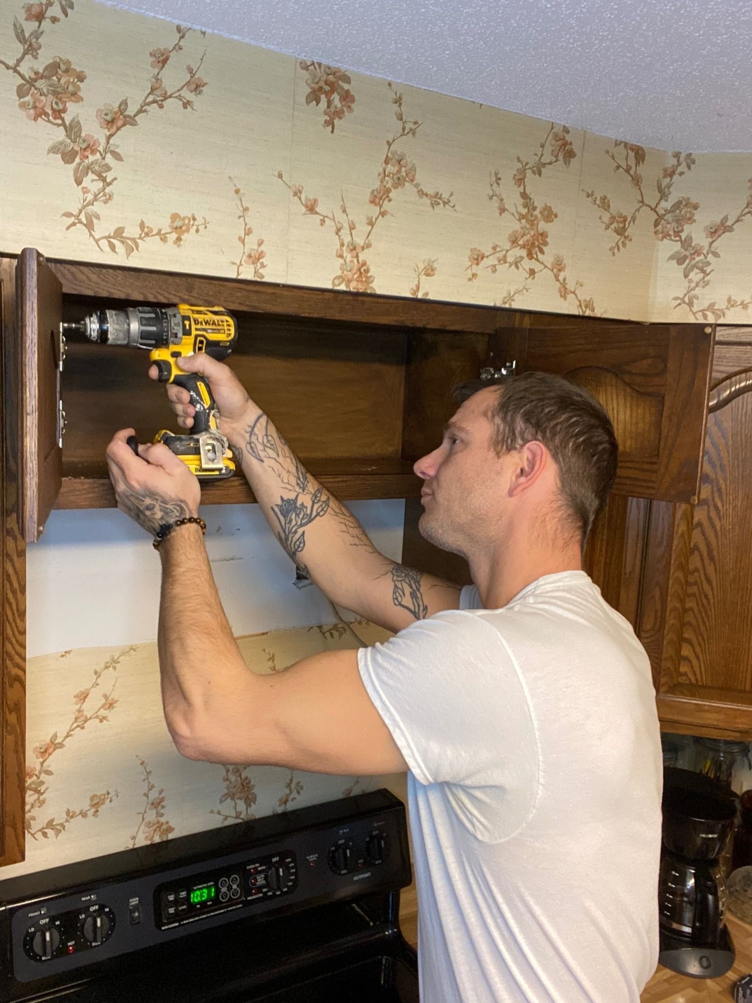 Man using a drill on a wooden cabinet in a kitchen with floral wallpaper.