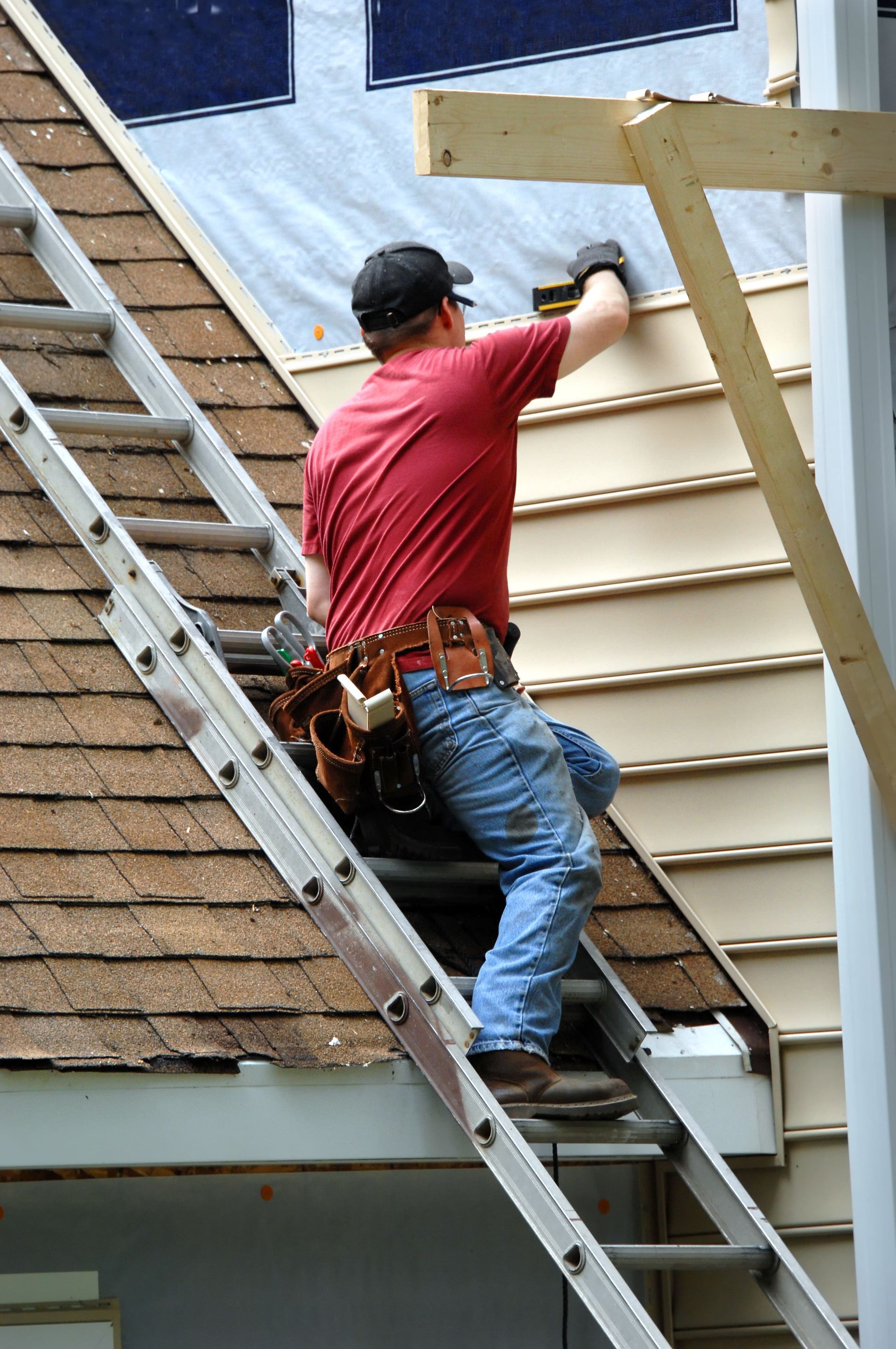 Man on ladder installing siding on a house. He wears a red shirt, blue jeans, and a tool belt.