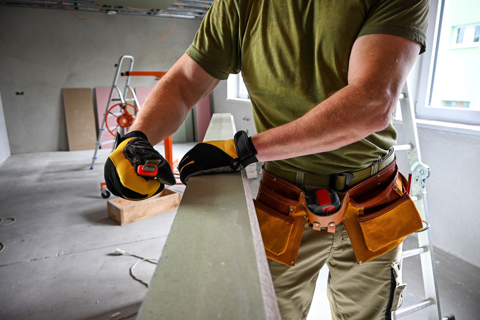 Construction worker sanding wood indoors, wearing gloves and tool belt.