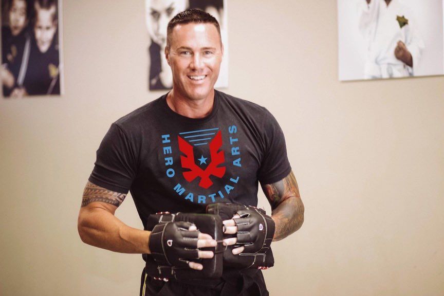 A man wearing a martial arts shirt is holding a pair of gloves.