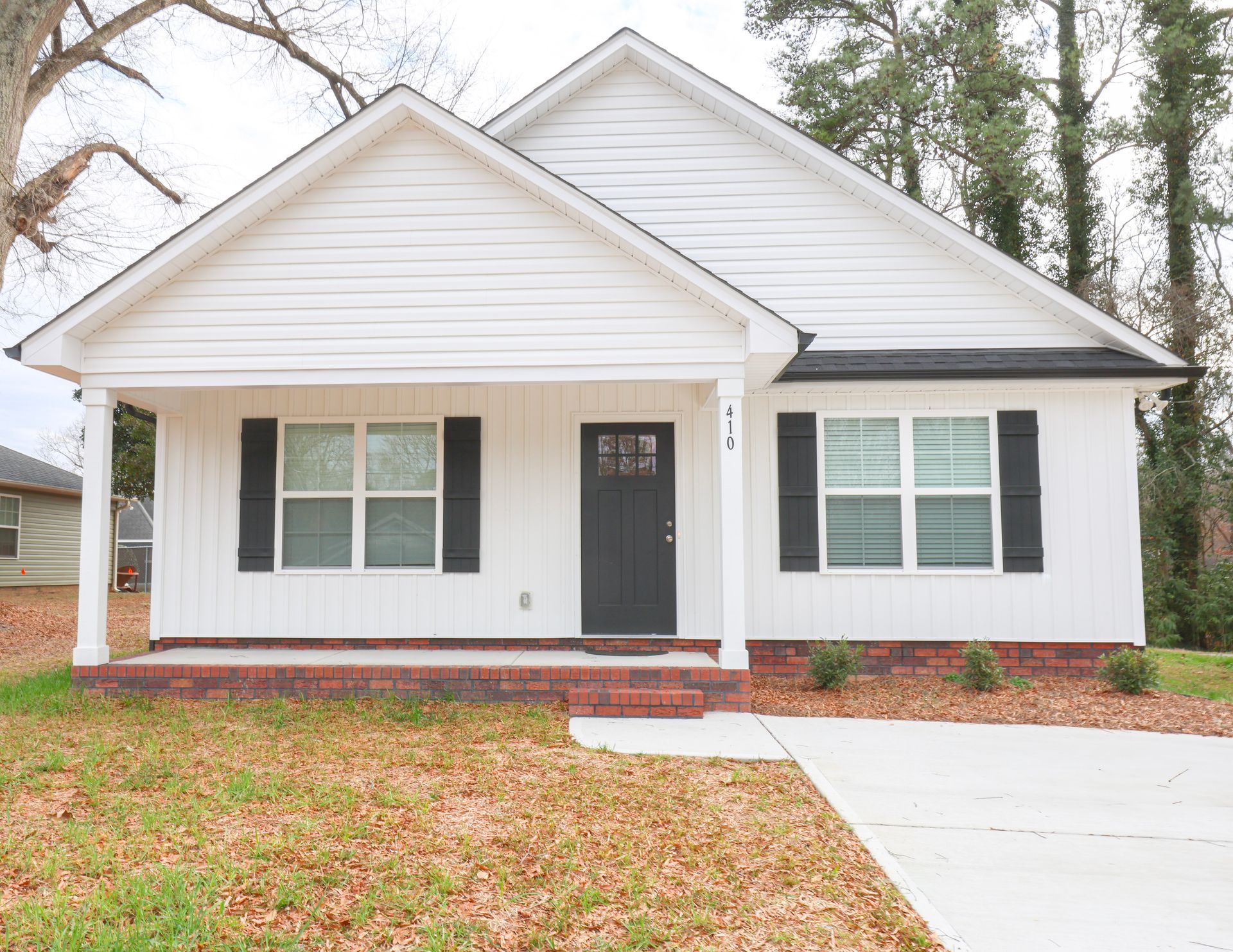 White house with black shutters, dark door, and brick foundation, with a concrete driveway. | Next Level Family Solutions