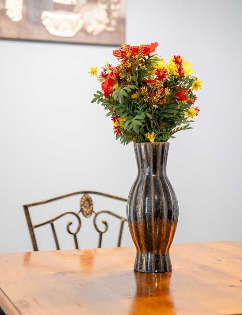 Flowers in a striped brown vase on a wooden table, with a chair and art in the background.