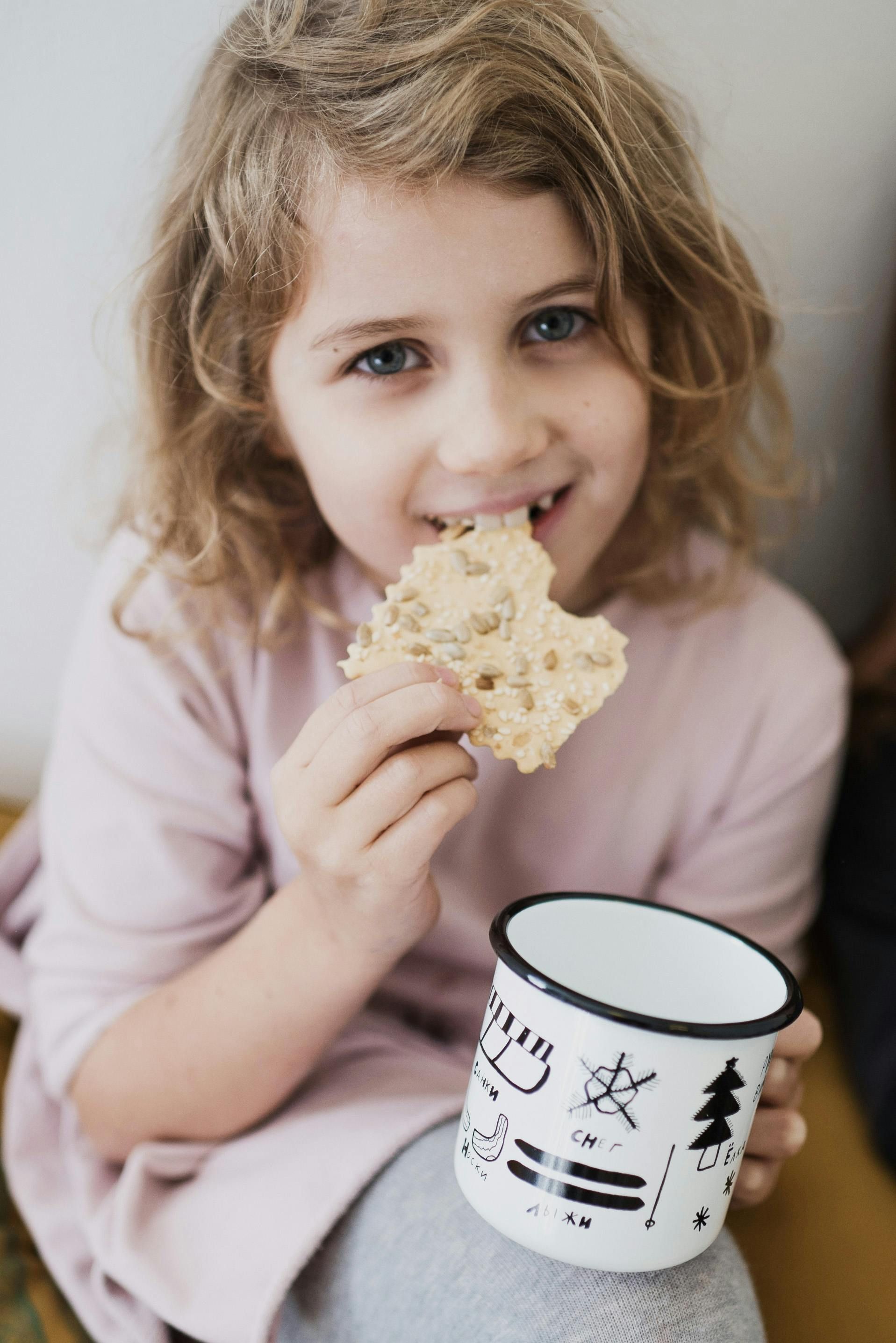 Young child with wavy blonde hair, eating a cracker and holding a white mug.
