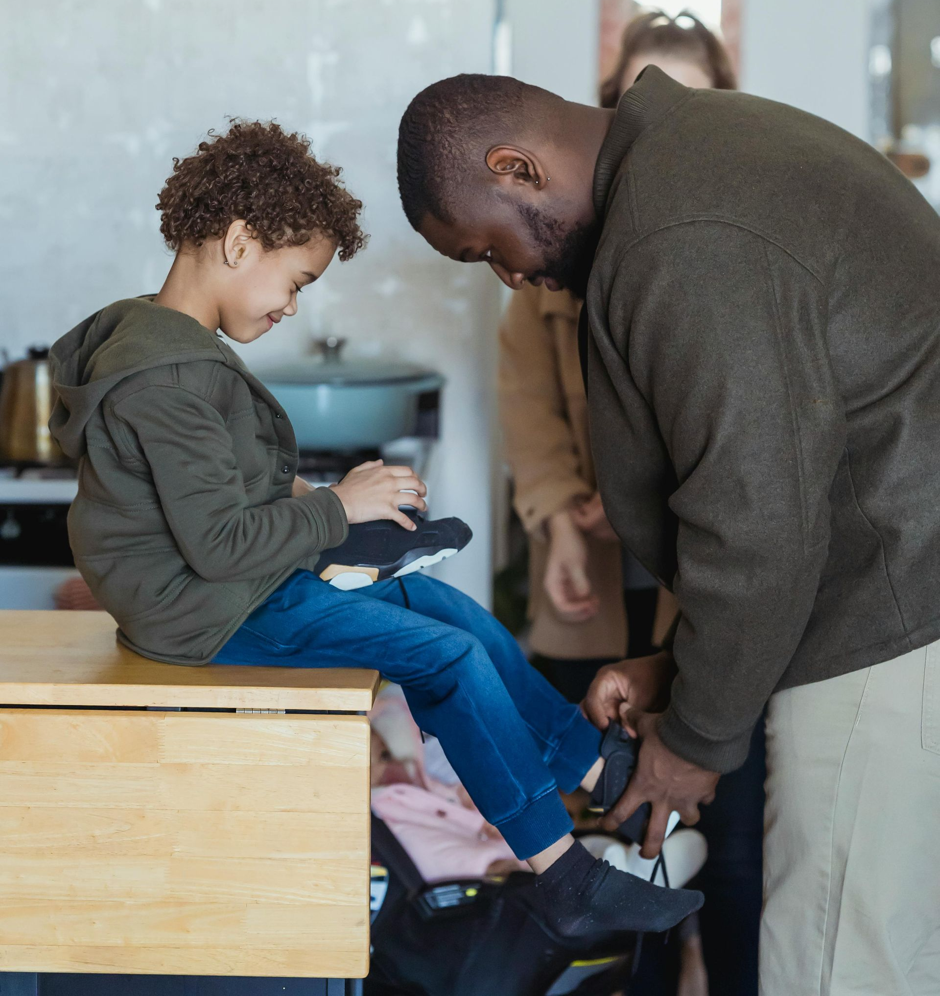 Man helping a child put on their shoes; indoor, kitchen.
