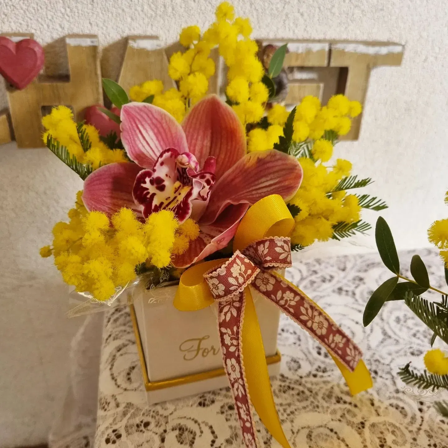 Un vaso pieno di fiori gialli e un fiore rosa con un nastro che dice