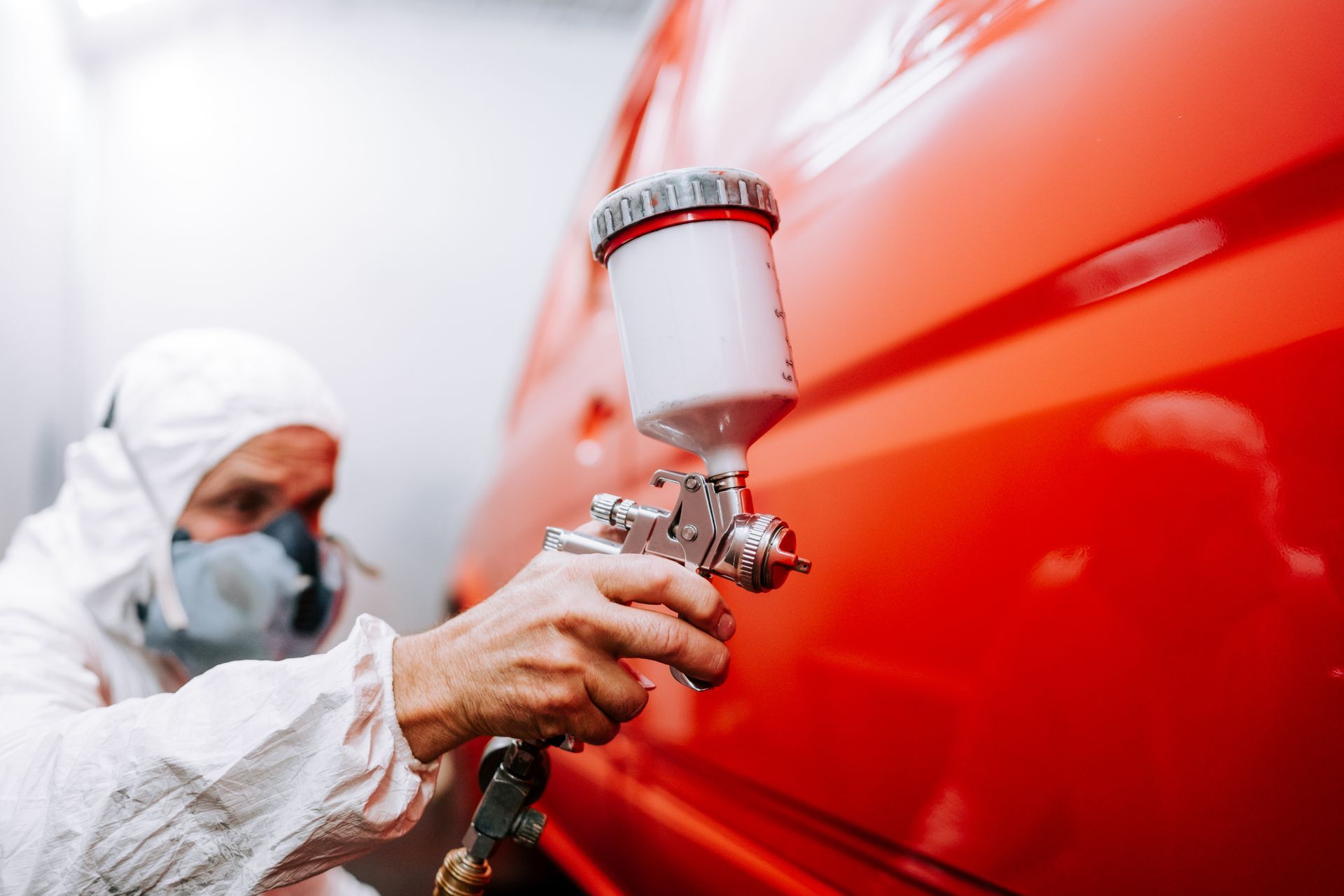 A car spray painter painting a car red in a painting box, wearing protective gear.