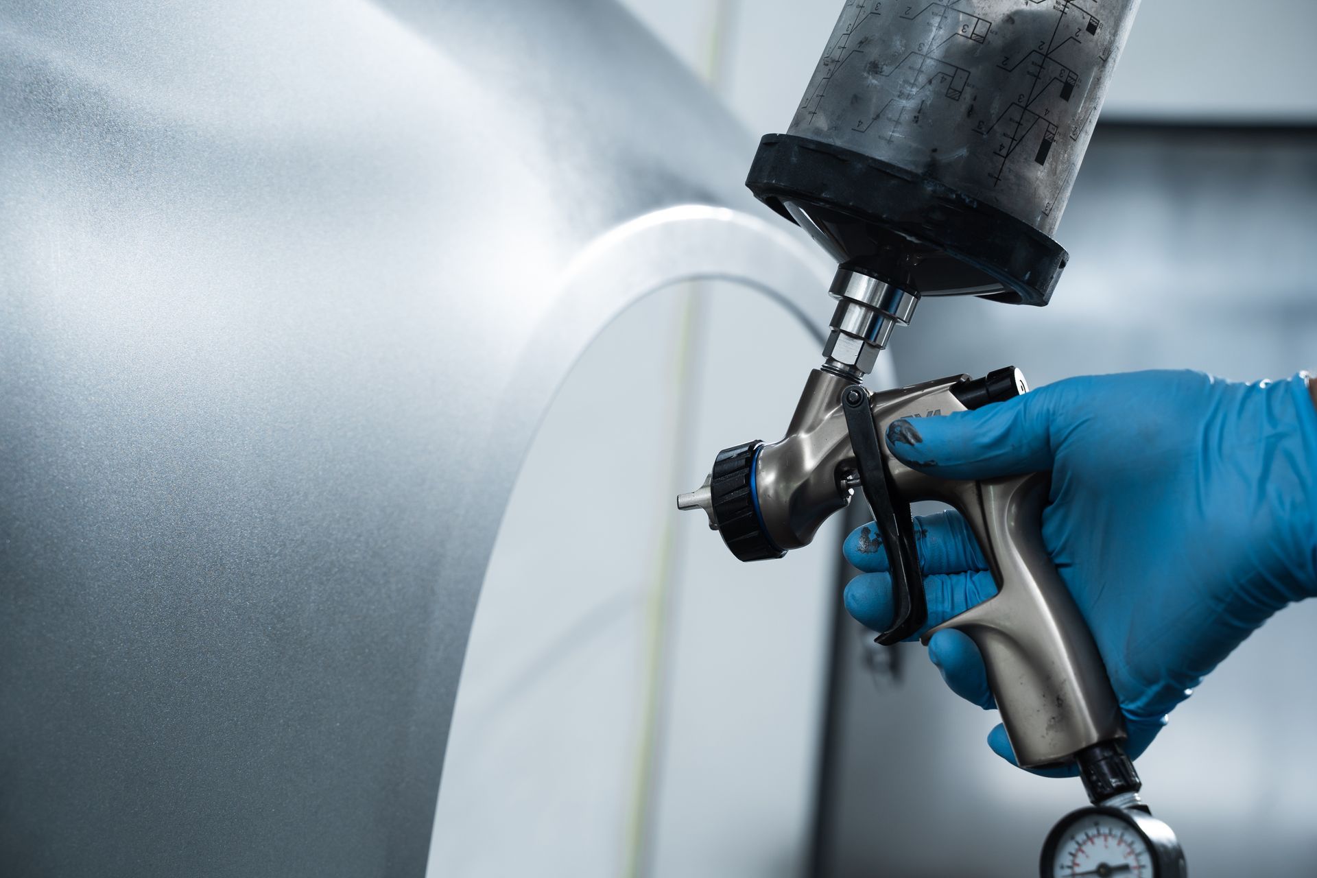 Person in blue gloves using a spray gun to paint a car panel silver in a workshop.