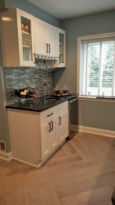 White mini-bar with black countertop, glass backsplash, and wine glasses hanging under cabinets.