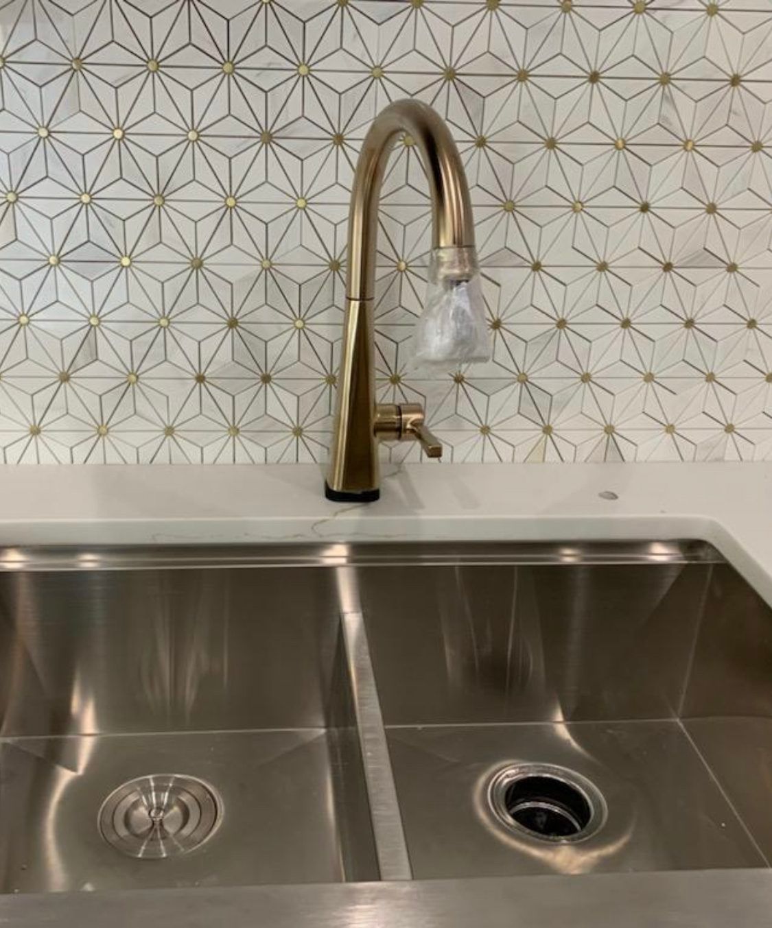 Gold faucet over stainless steel double sink, with geometric patterned backsplash.
