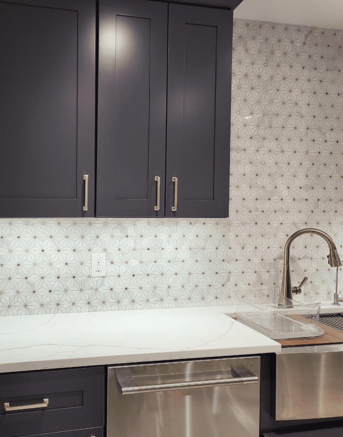 Dark blue kitchen cabinets above a white countertop, stainless steel sink and dishwasher, and geometric backsplash.