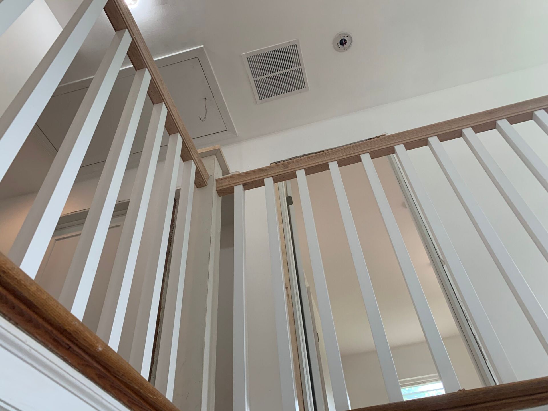 Looking up at a white staircase with wooden handrails, ventilation, and a hatch in a bright room.