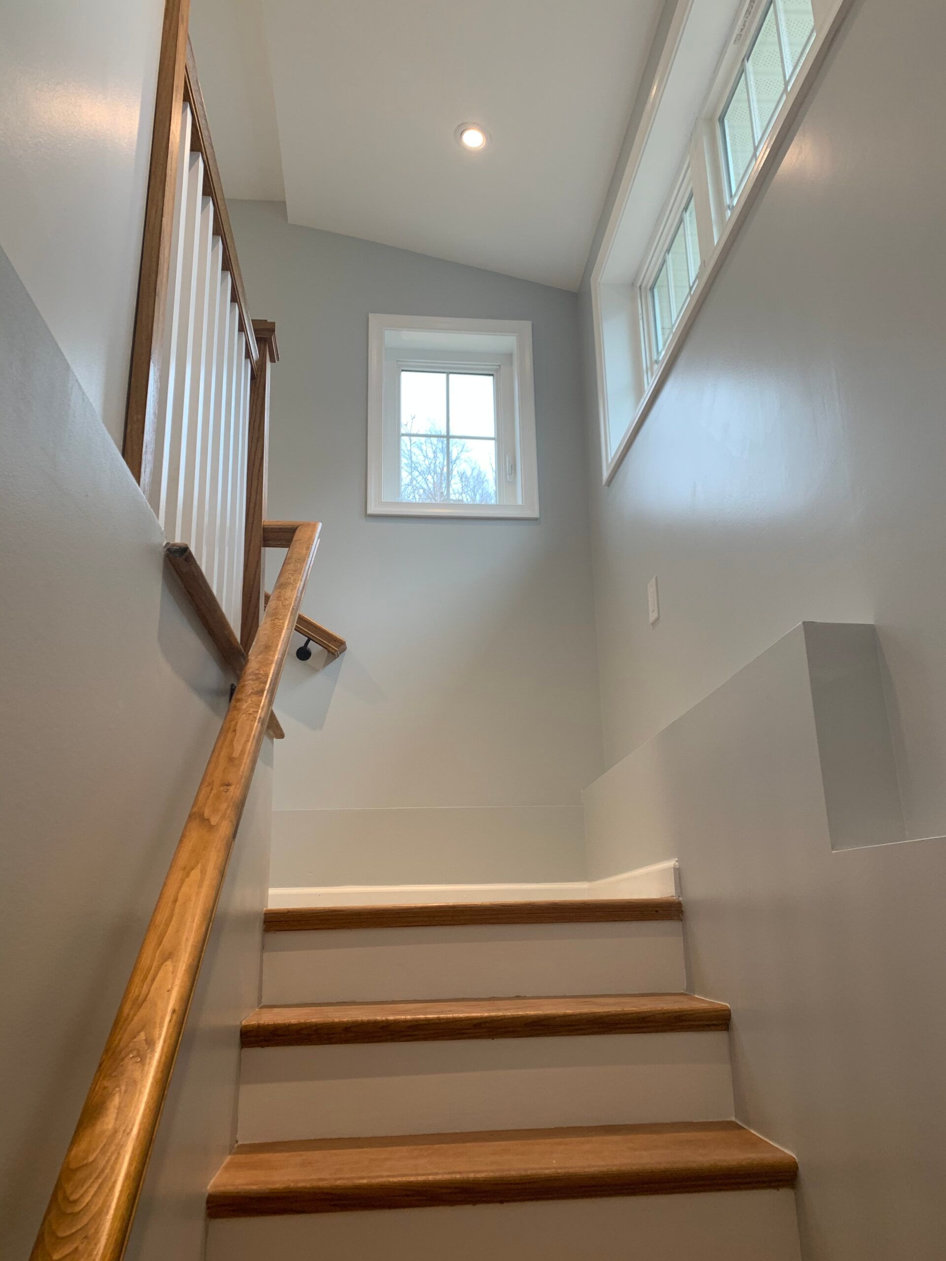 Staircase with wood handrail and light gray walls; small windows above.