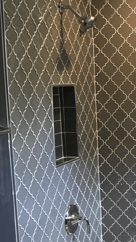 Shower interior with gray and white patterned tiles, built-in shelf, and silver fixtures.