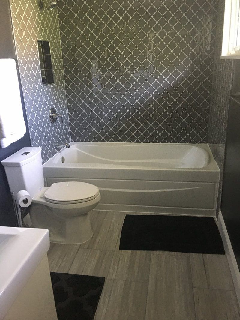 Bathroom with white toilet, tub, and gray patterned wall tiles, plus black bath mats.