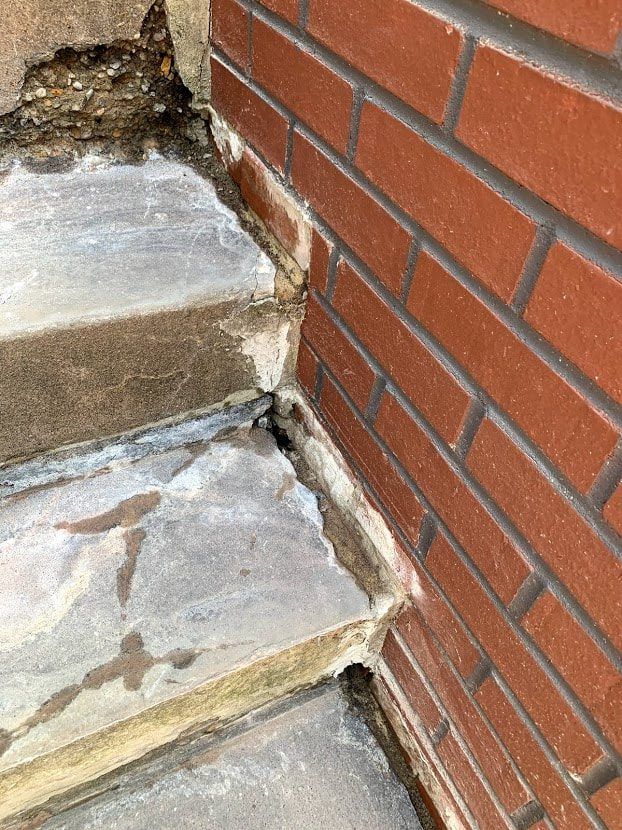 Brick wall corner meets stone steps, showing crumbling mortar and uneven surfaces.