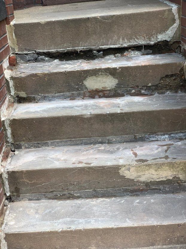 Concrete steps, worn and damaged, leading upwards against a brick wall.