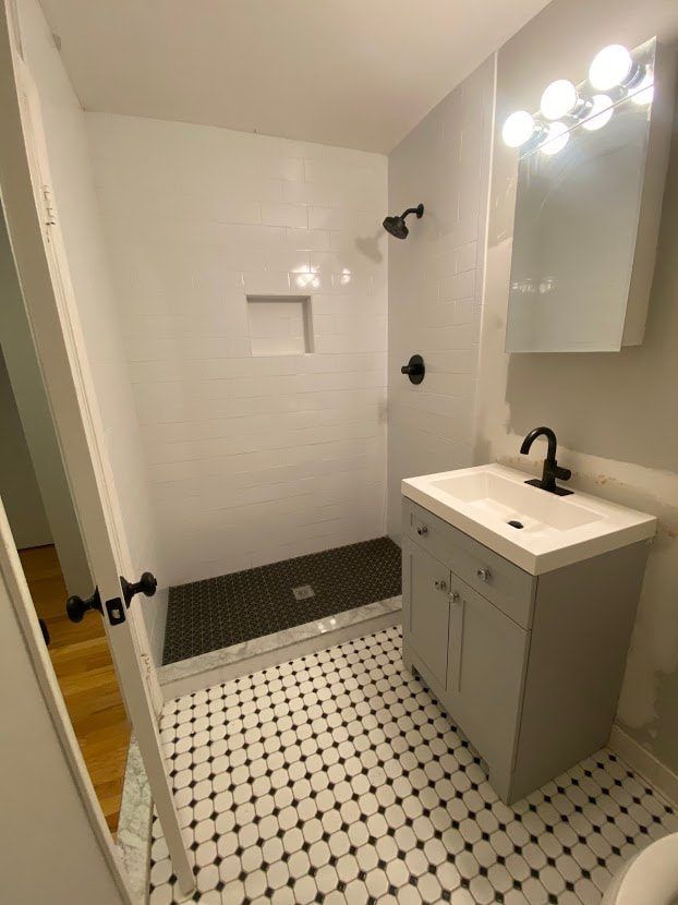 Bathroom with white tiled shower, black fixtures, gray vanity, and black and white patterned floor.