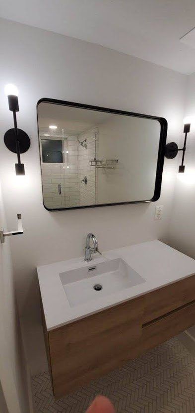 Bathroom vanity with a rectangular black-framed mirror, two sconces, a white sink, and a wooden cabinet.
