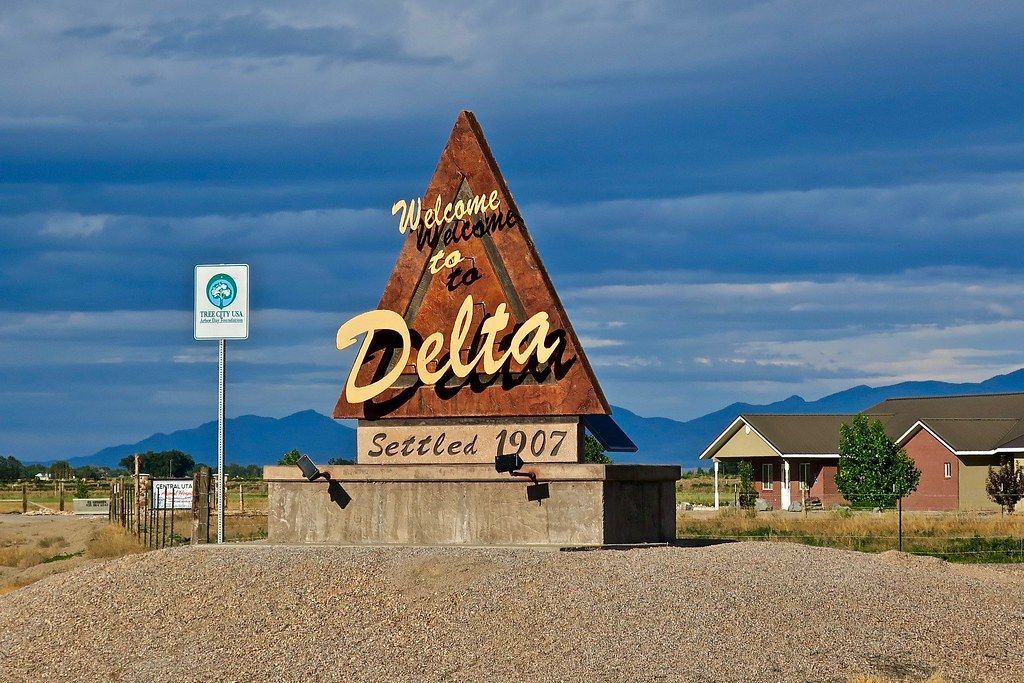 A triangular, rust-colored sign welcoming visitors to Delta, standing on a gravel mound against a cloudy sky.