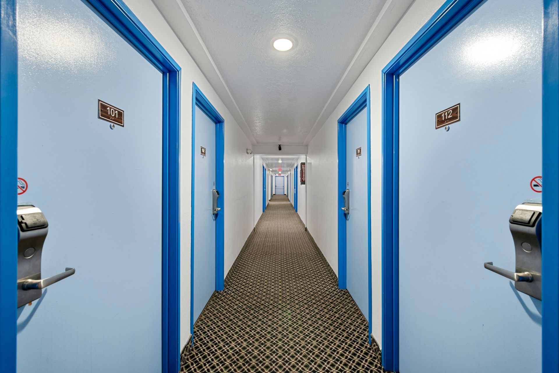 A long hotel hallway with pale blue doors, patterned brown carpet, and bright overhead lights.