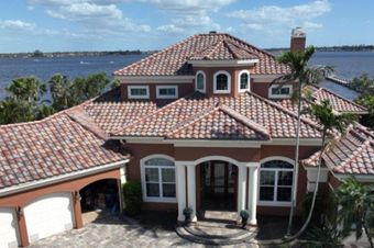 A house with a dark gray shingle roof and a tan exterior. Other houses are in the background.