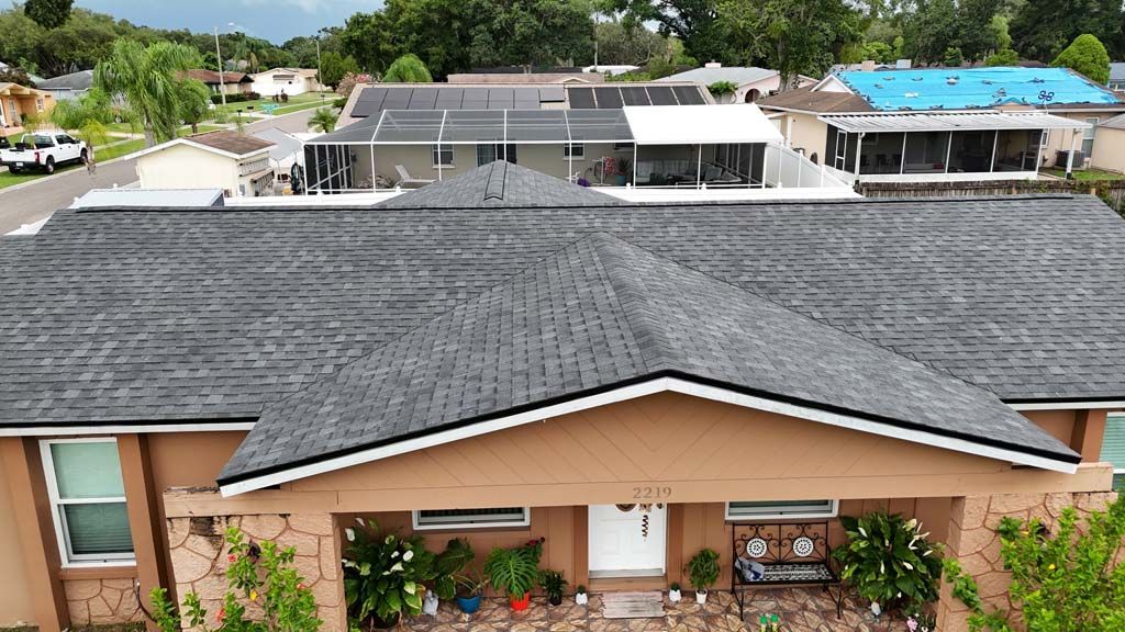 A house with a dark gray shingle roof and a tan exterior. Other houses are in the background.